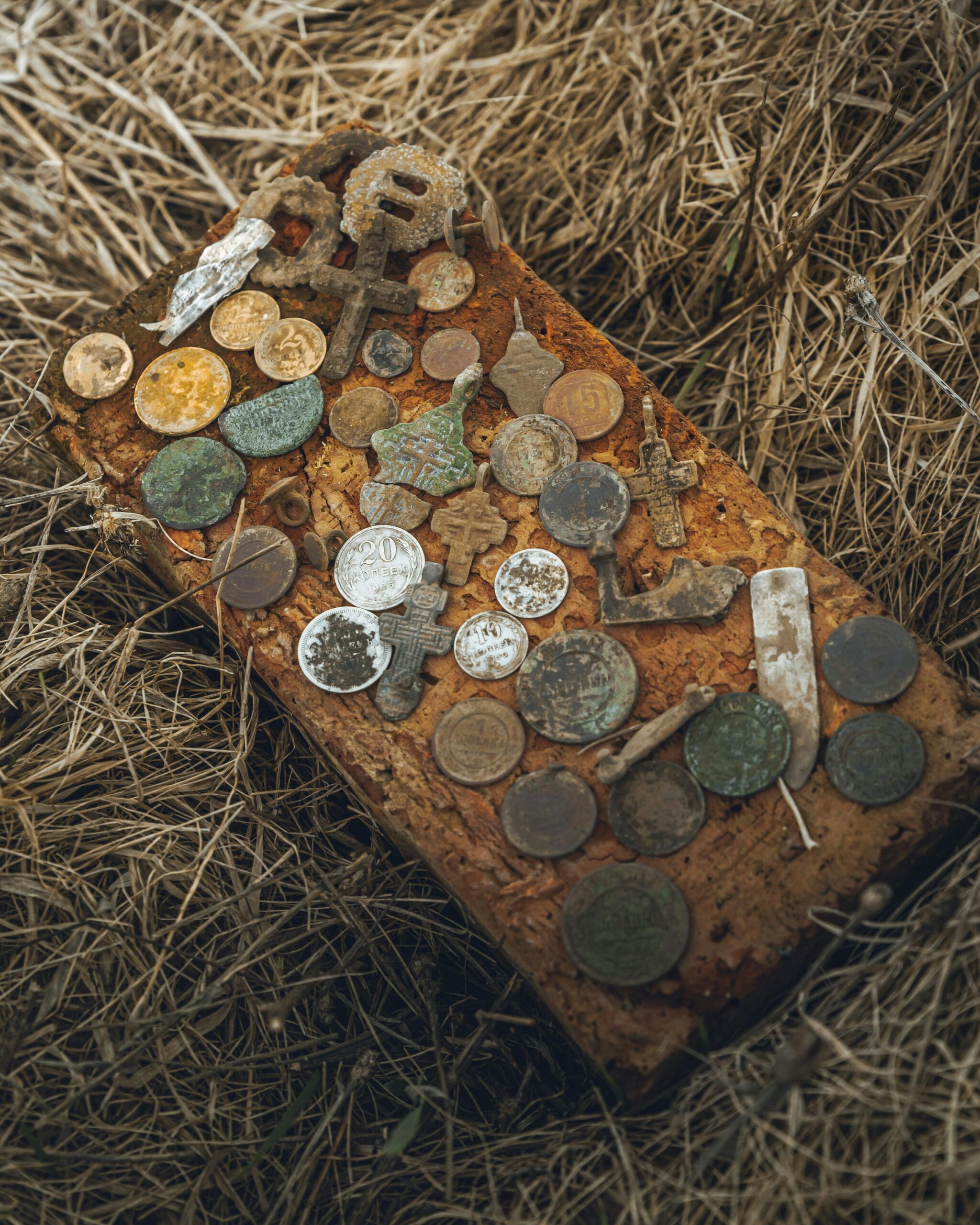 Collection of old coins and artifacts on a rusted surface amid straw, capturing historical essence.