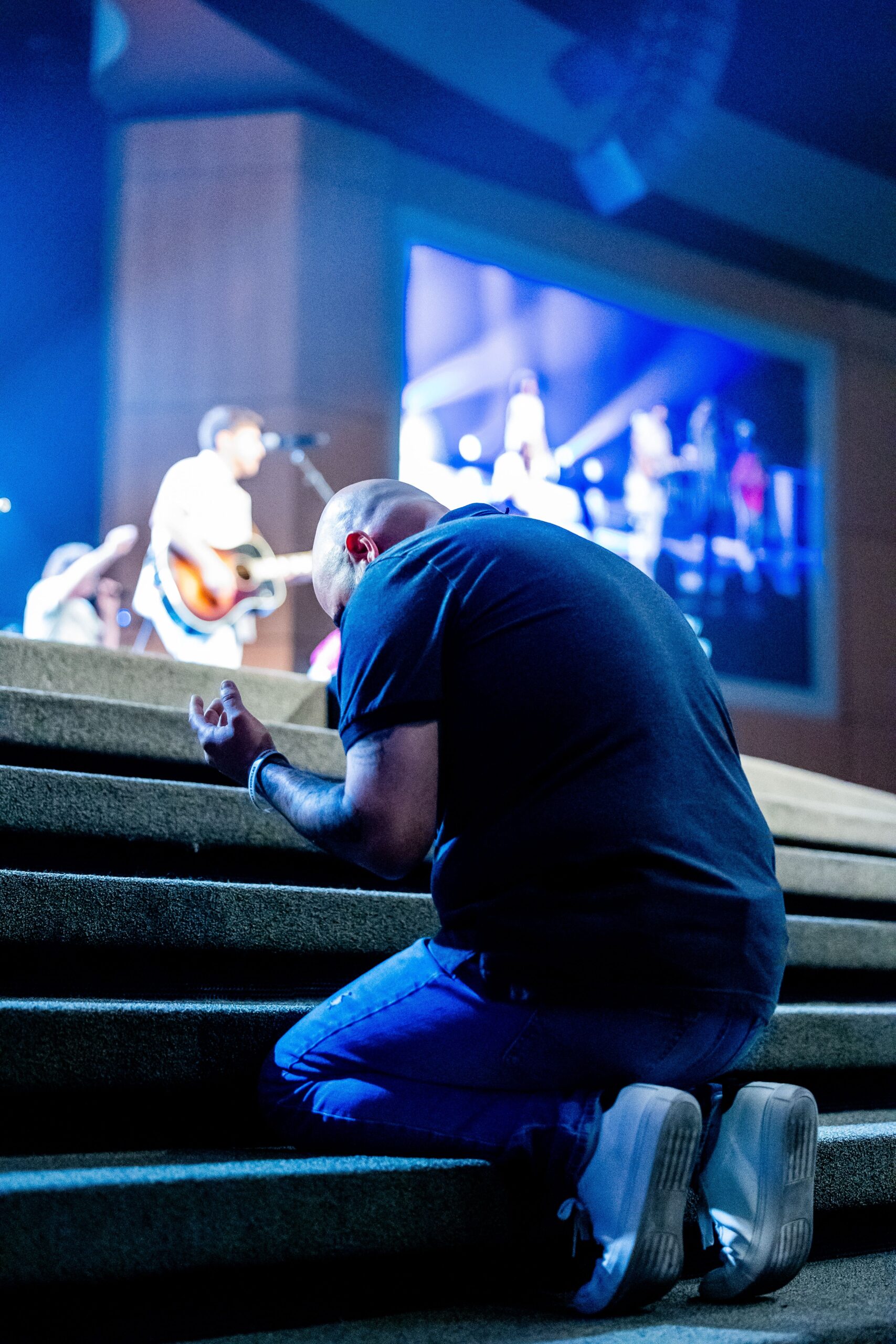 A person kneels in prayer at a church service, while a live band performs on stage.